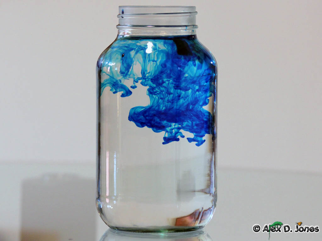 A glass jar filled with clear water shows bright blue liquid spreading and swirling through it.