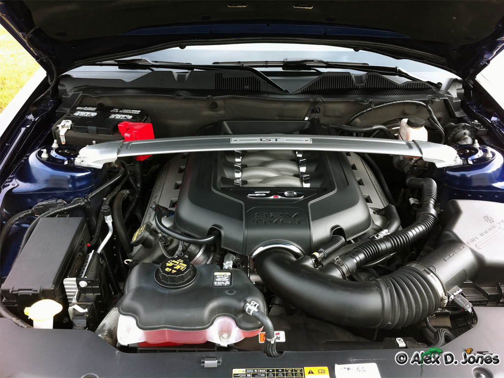 This picture shows the inside of a car engine with its hood open. You can see many metal parts, tubes, and wires that work together to make the car move.