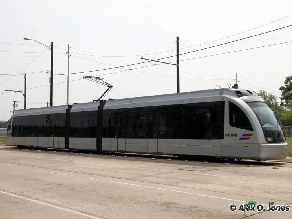 This picture shows a long, white train called a light rail that runs on tracks through a city.
