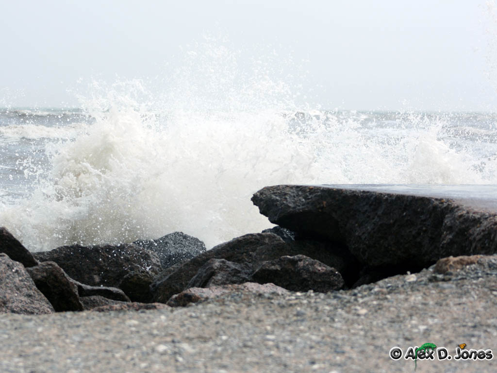 Ocean waves crash against dark rocks on a beach, creating white foam and spray. The water hits the rocks with great force, sending water droplets high into the air.
