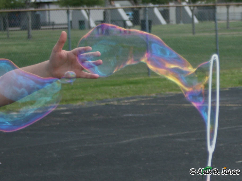 A person is reaching toward a large, colorful soap bubble floating in the air. The bubble shows rainbow colors like purple, blue, green, and yellow on its thin surface.