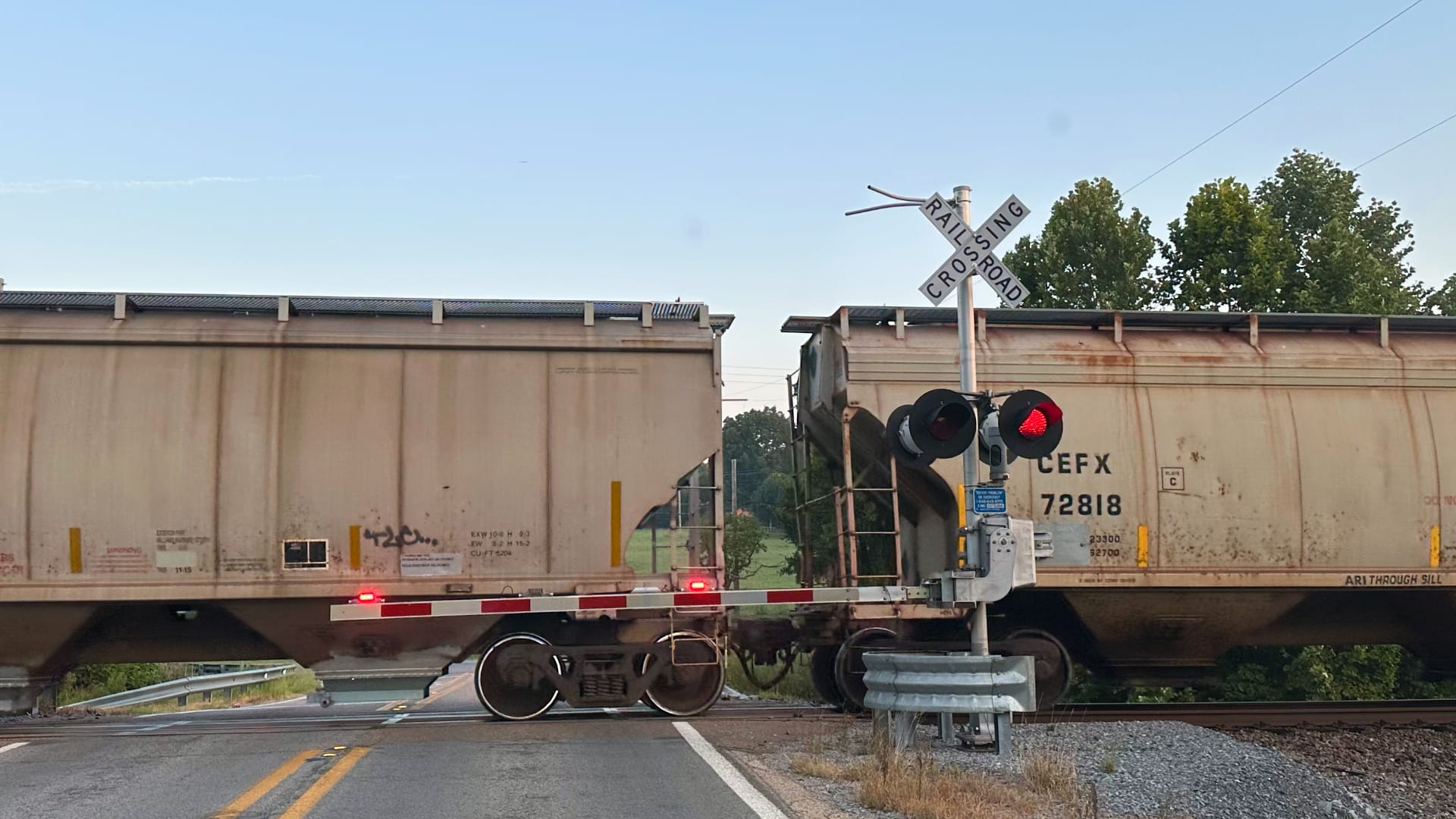 The photo shows a railroad crossing with a train stopped. Two large train cars are visible, with a railroad crossing sign and flashing red lights indicating that a train is present.
