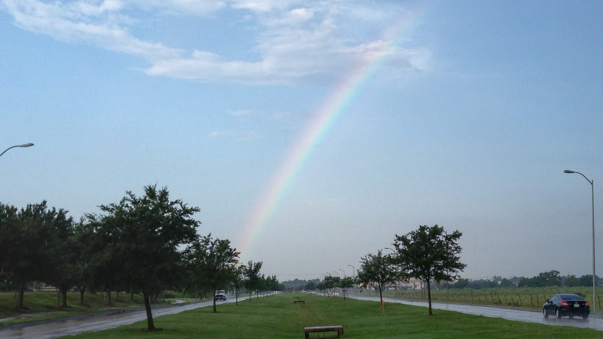 This image shows a beautiful rainbow stretching across a rainy sky above a divided highway with green grass, trees, and street lights on both sides.