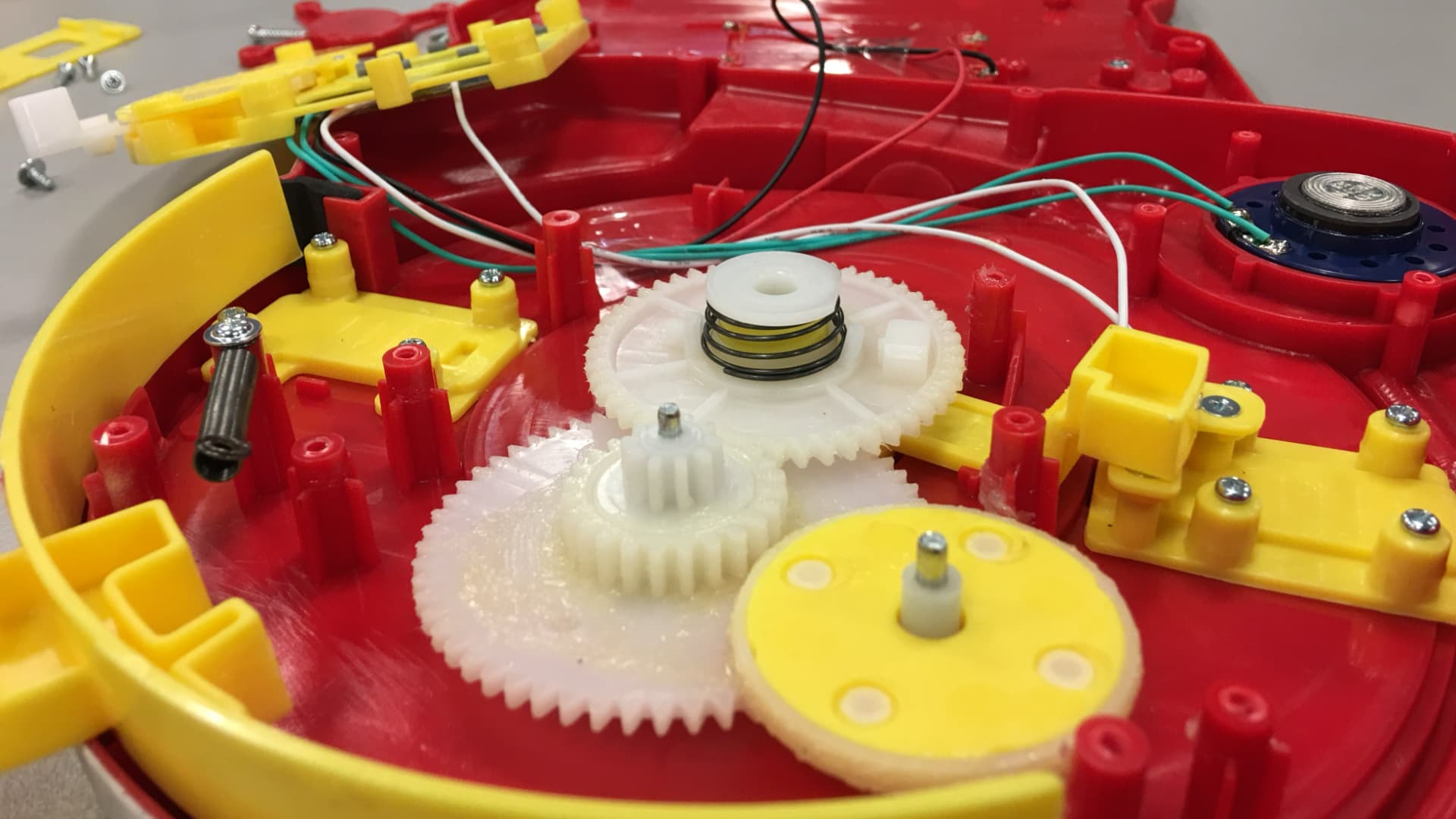 This image shows the inside of a disassembled toy. Several plastic gears of different sizes are visible, along with wires, a small speaker, and plastic pieces that hold other components in place.
