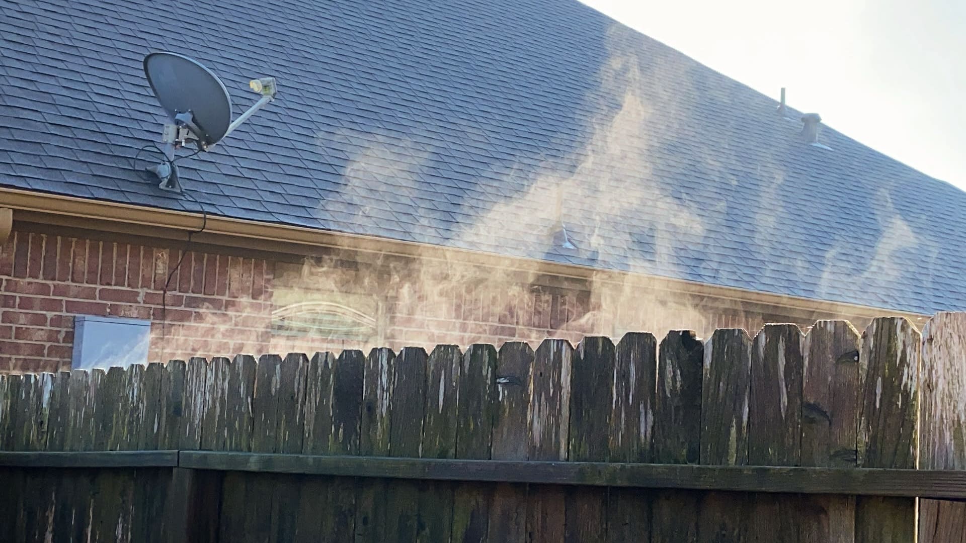 This photograph shows steam rising from a chimney on a brick building with a dark roof on a clear day.