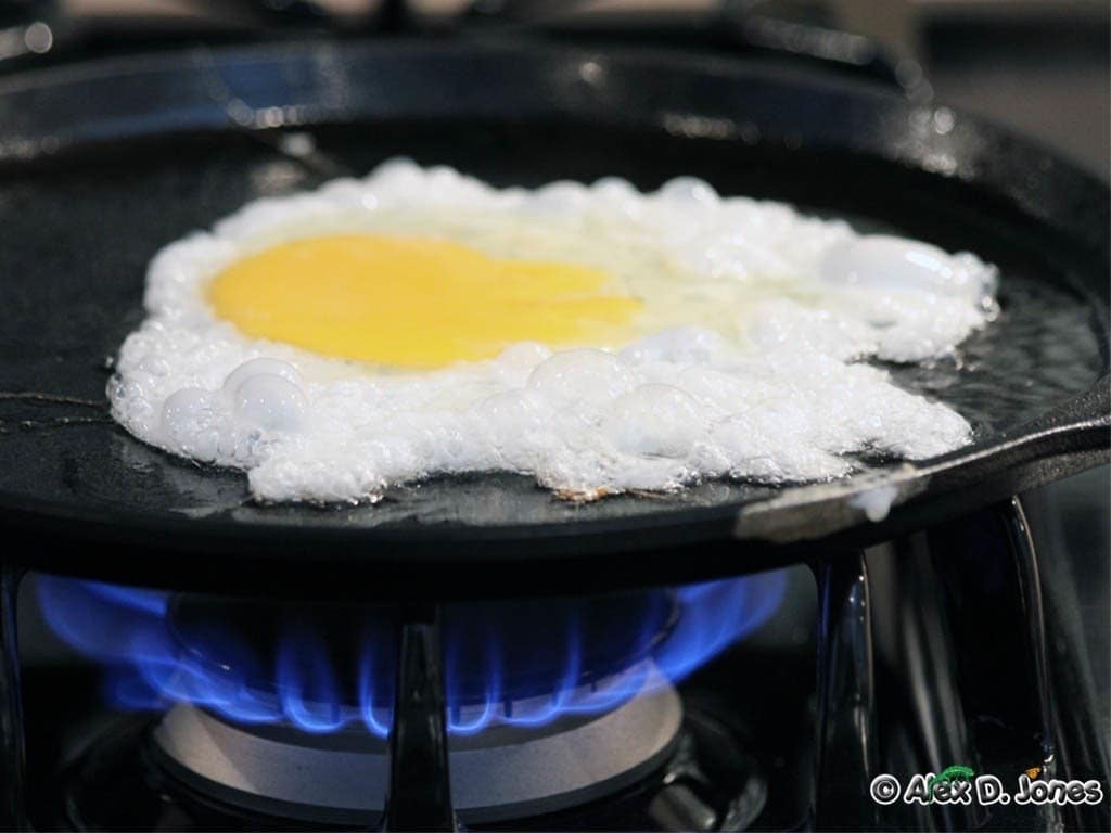 This image shows a fried egg cooking on a dark skillet over a blue gas flame. The egg white has turned from clear and runny to white and solid, while the yellow yolk in the center remains soft.