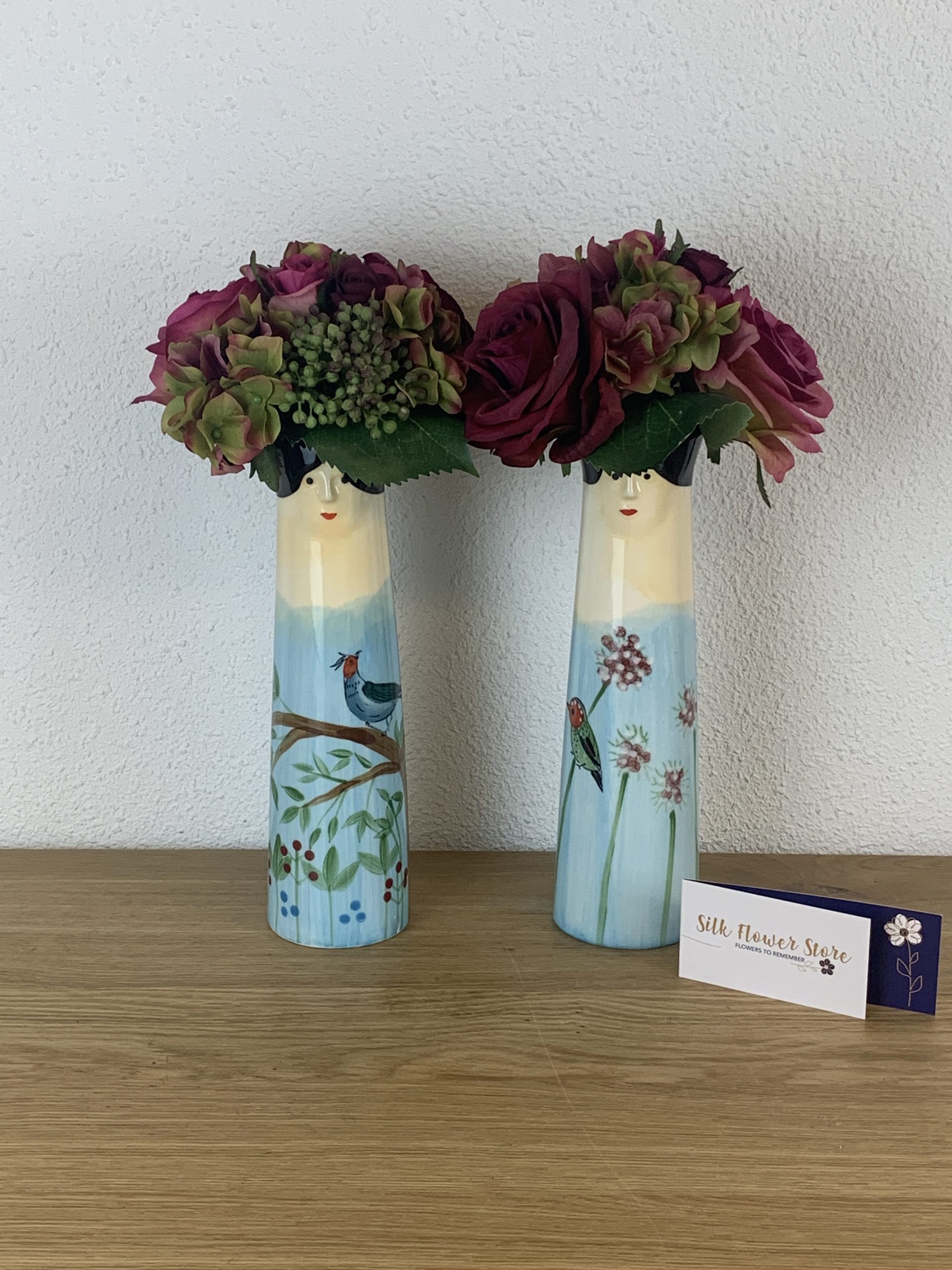Twee keramische vazen in de vorm van damesfiguren, gevuld met dieprode zijden bloemen, staand op een houten ondergrond.