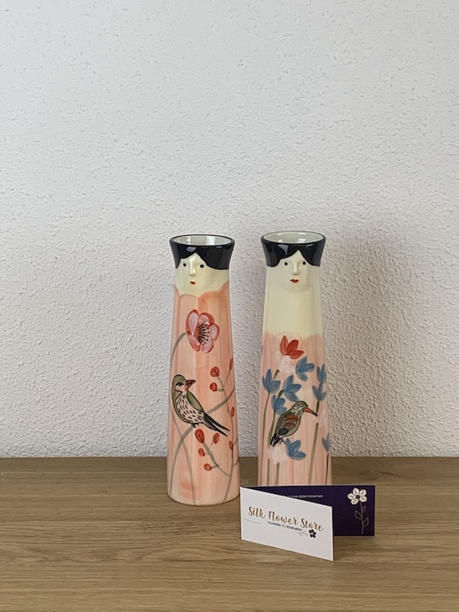 Twee keramieken vazen met een vrouwenhoofd, versierd met vogels en roze bloemen, naast een visitekaartje.