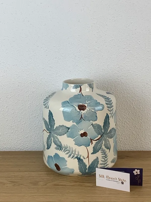 Een roomwitte keramische vaas met een aquarelachtig patroon van blauwe bloemen en bladeren op een houten ondergrond.