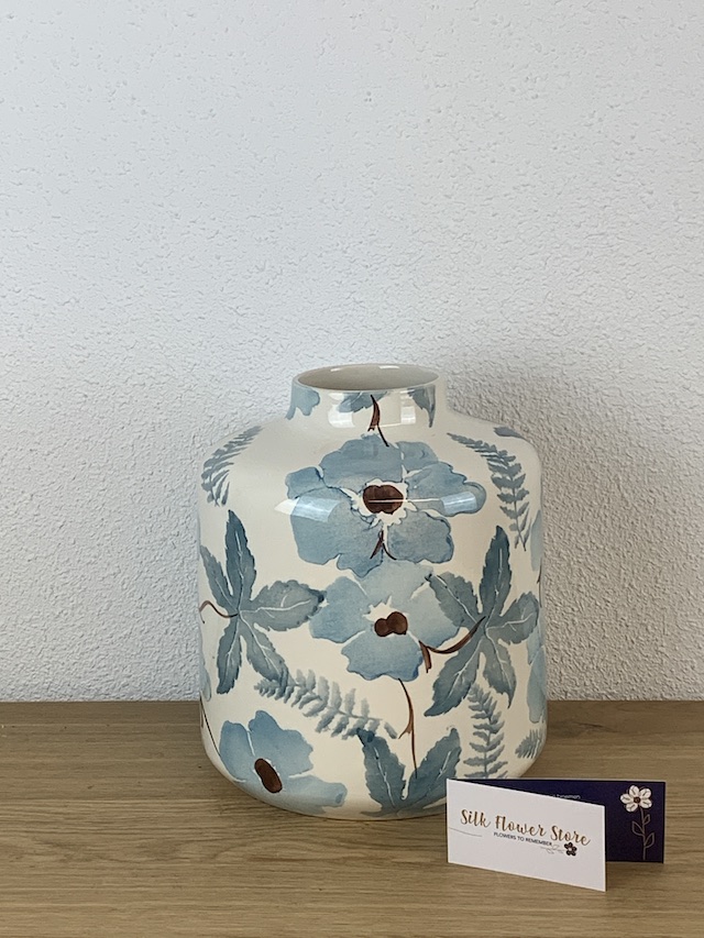 Een roomwitte keramische vaas met een aquarelachtig patroon van blauwe bloemen en bladeren op een houten ondergrond.