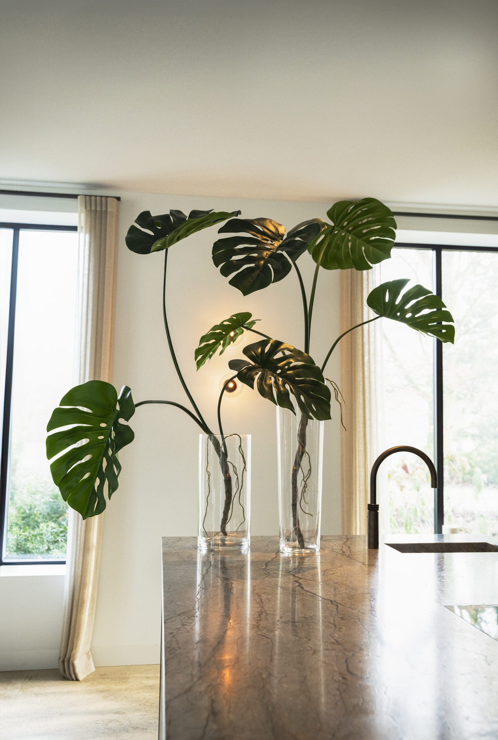 Twee grote kunstmatige Monstera-planten in glazen vazen op een marmeren keukeneiland in een modern interieur.