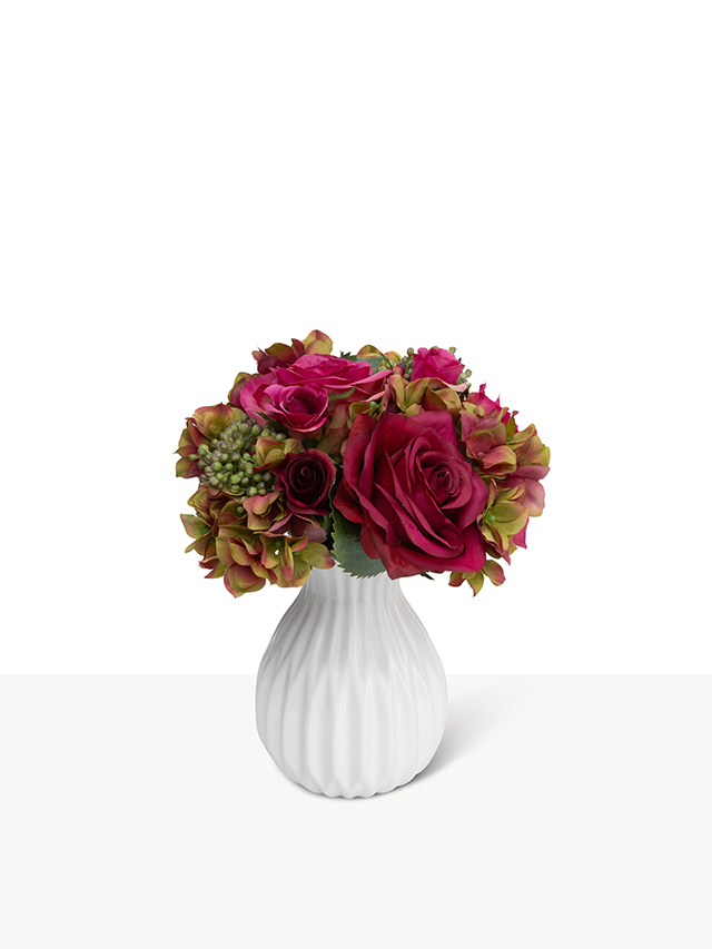 Een elegant mini boeketje van zijden bloemen in bordeauxrode en groenachtige tinten in een witte, geribbelde vaas.