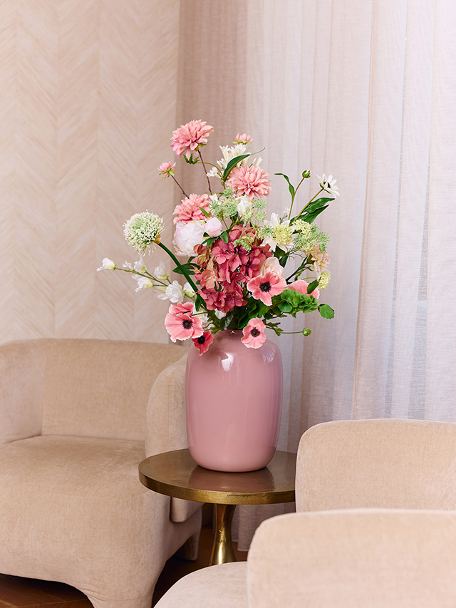Rijk zijden boeket met roze en witte bloemen in een roze vaas op een tafeltje in een licht interieur.