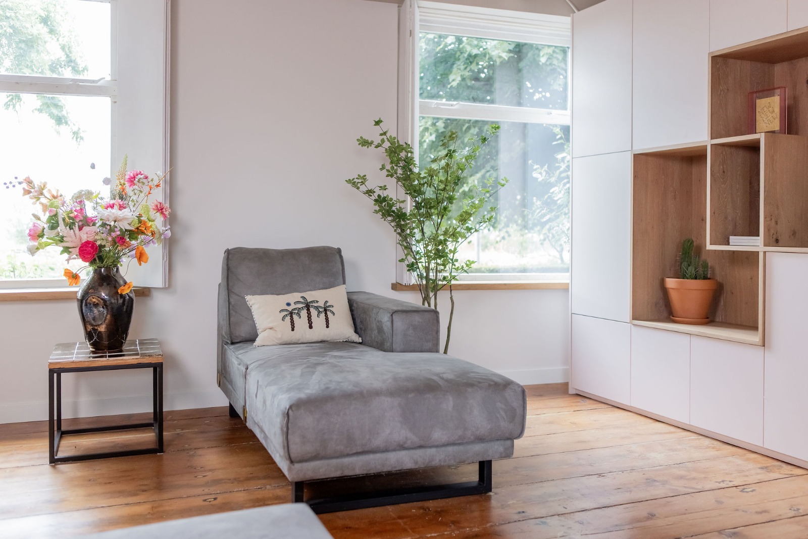 Kleurrijk zijden boeket in donkere vaas op bijzettafel naast grijze relaxstoel in lichte woonkamer.
