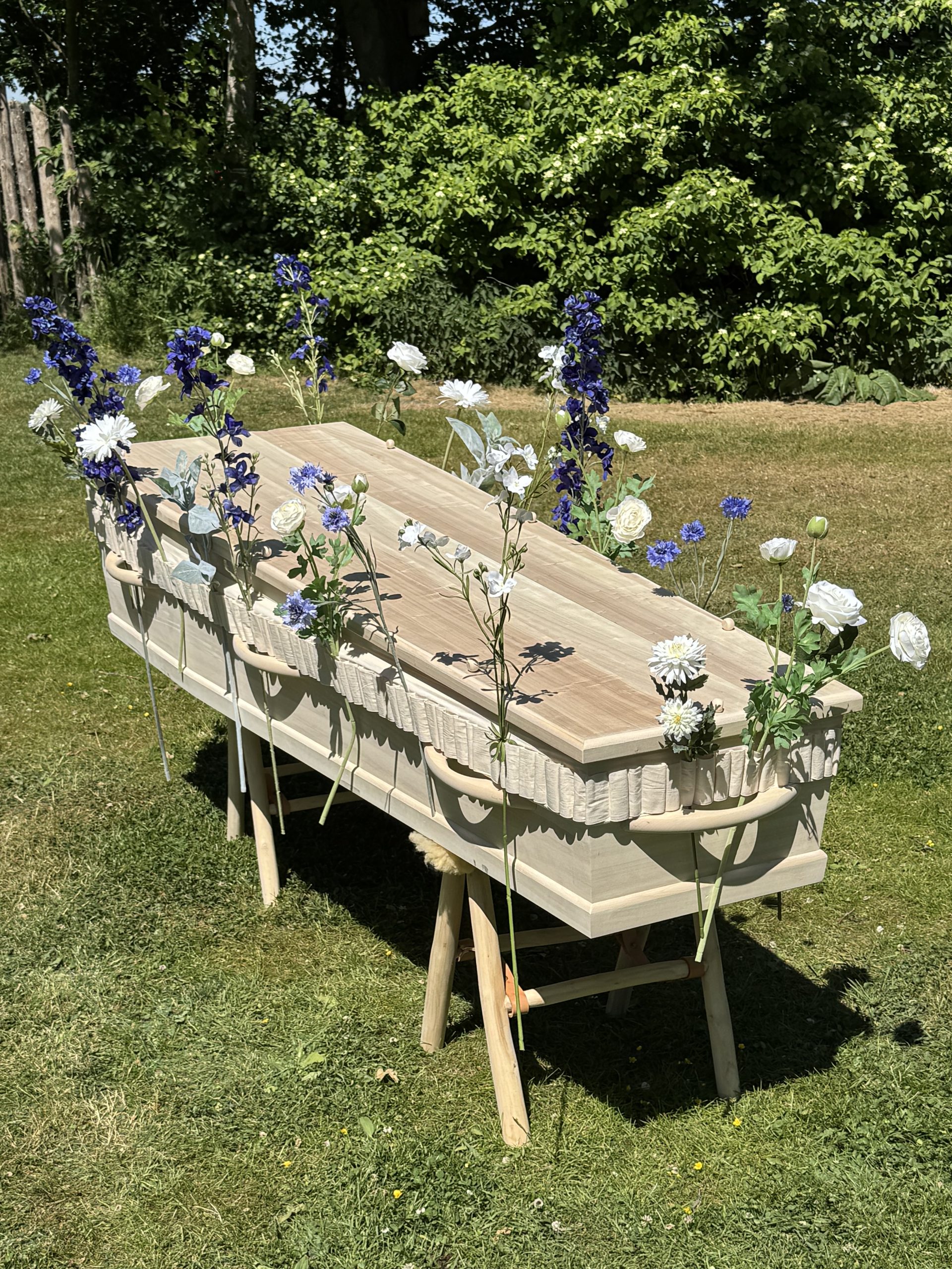Lichte houten kist op schragen versierd met blauwe en witte kunstbloemen in een groene tuin.