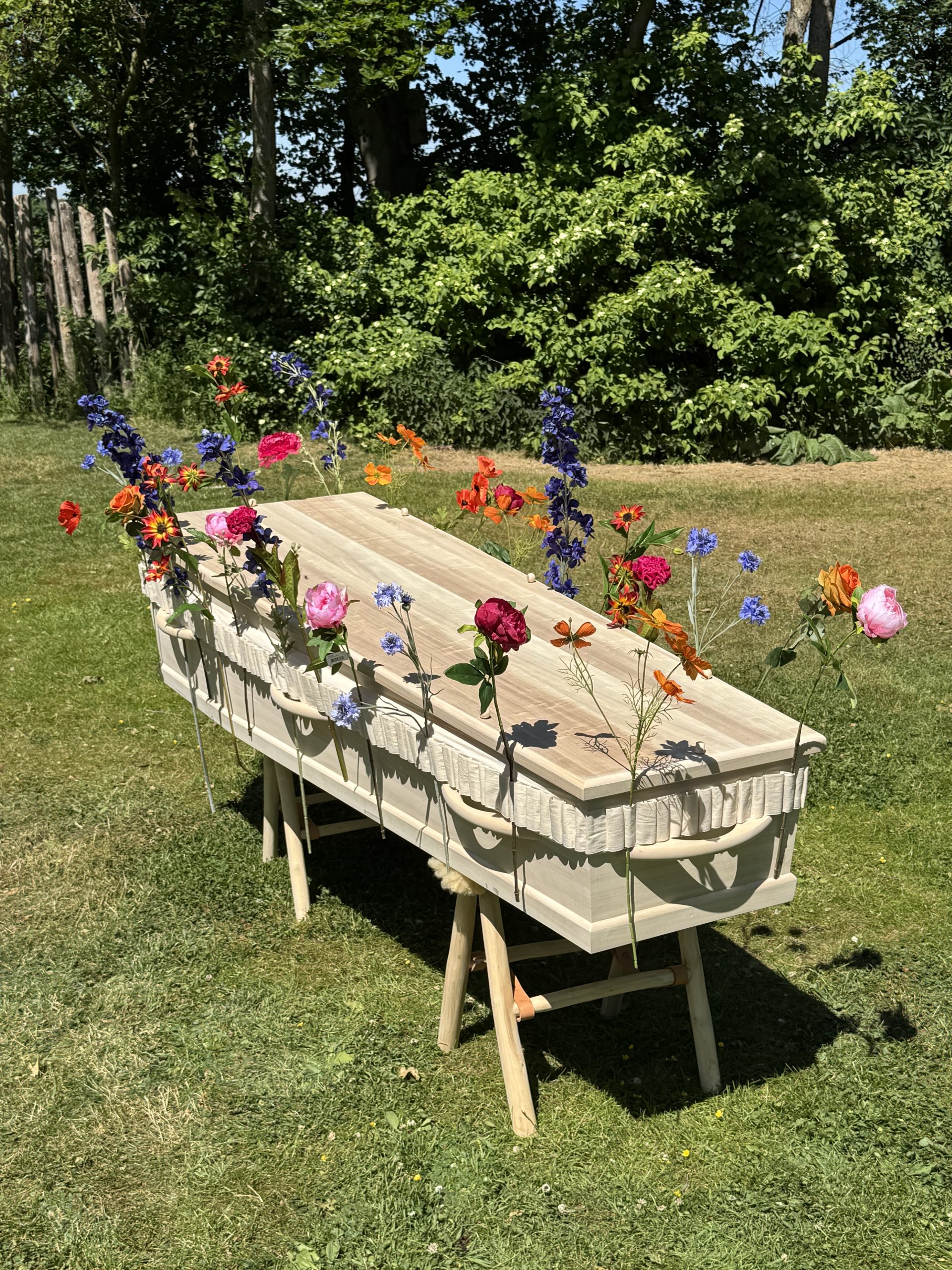 Een lichtgekleurde houten uitvaartkist versierd met kleurrijke zijden bloemen in een buitenomgeving.