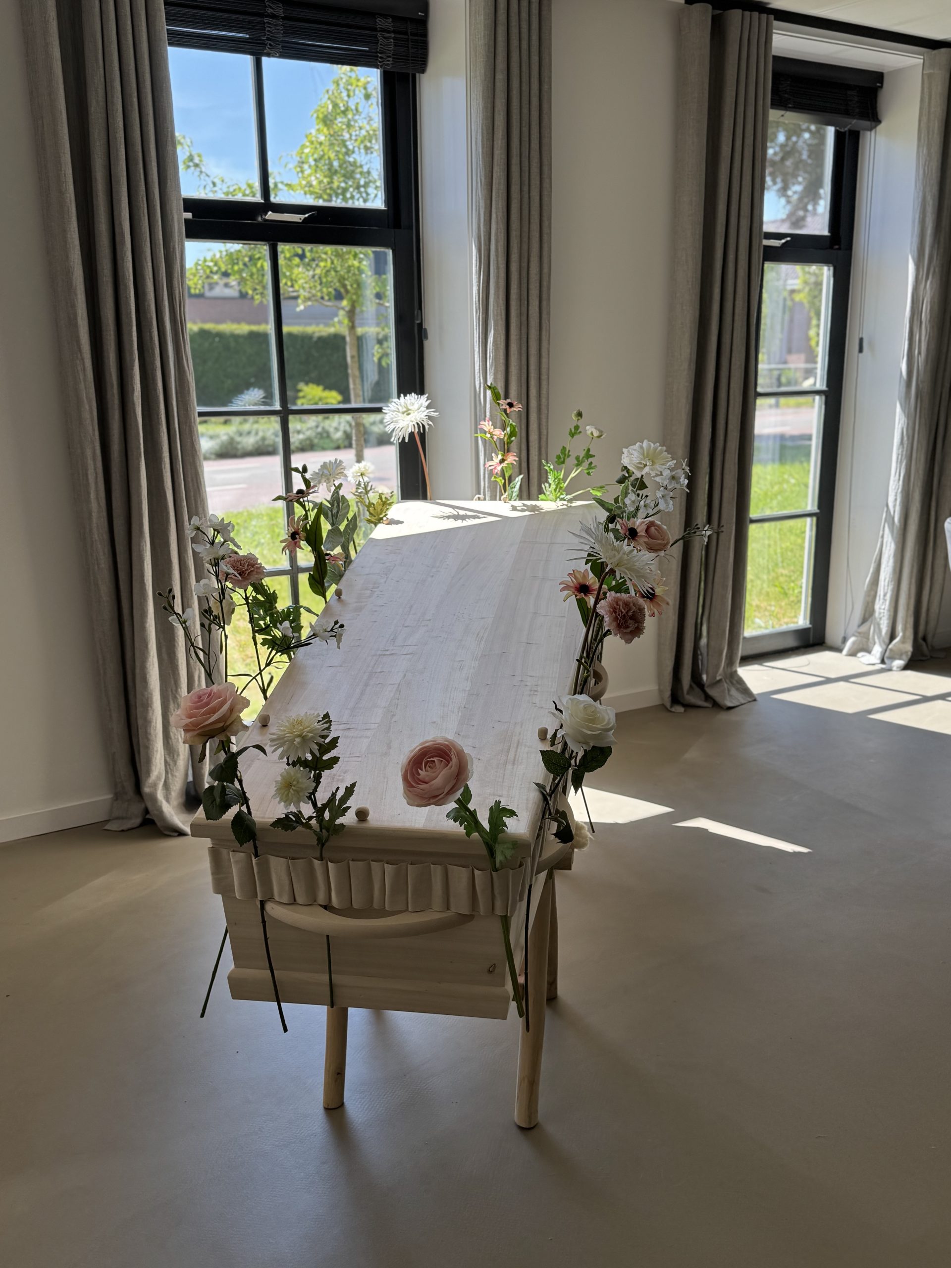 Licht houten rouwkist versierd met zijden bloemen in wit en zachte roze tinten in een elegant interieur.