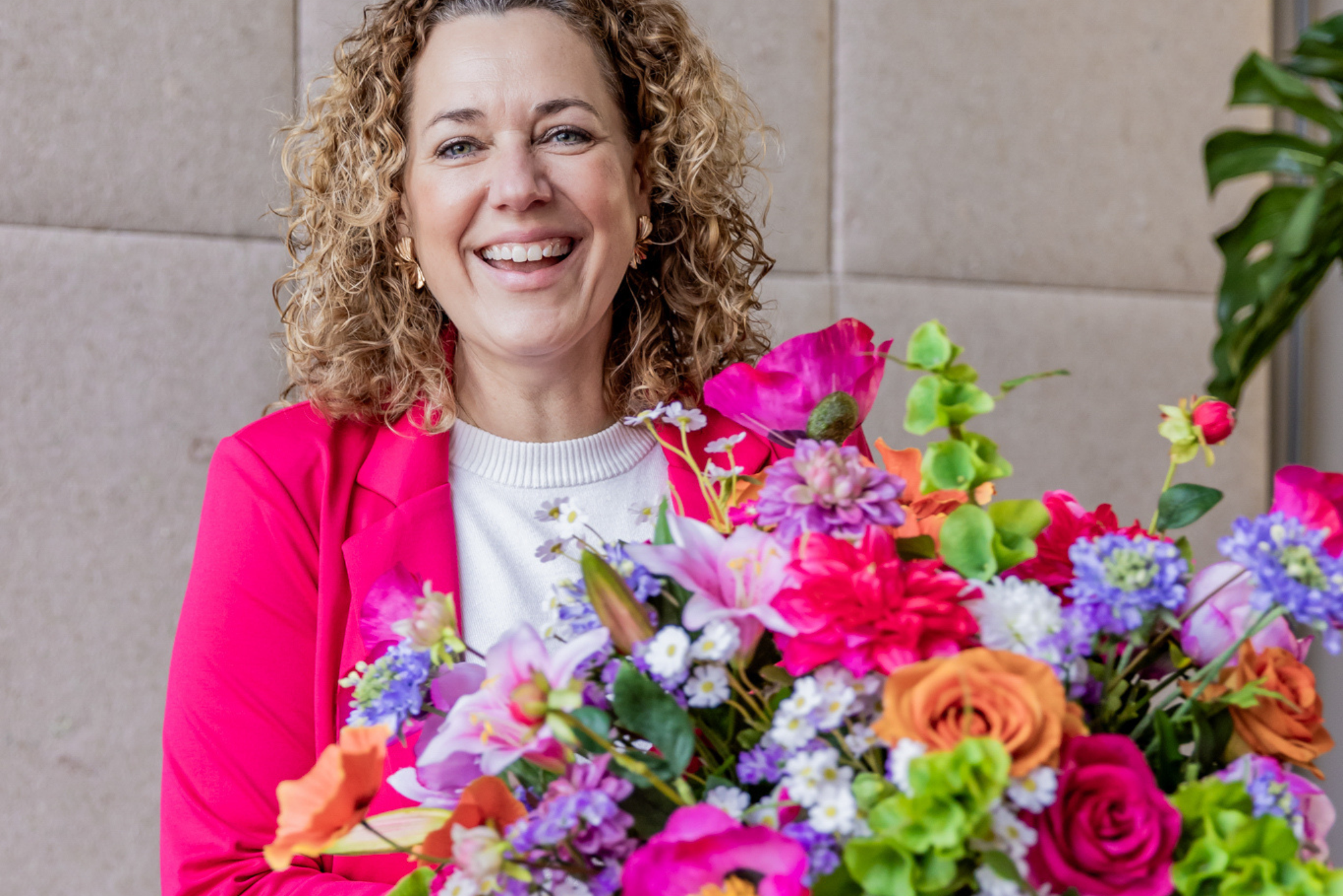 Lachende vrouw in een felroze blazer die een kleurrijk boeket zijden bloemen vasthoudt.