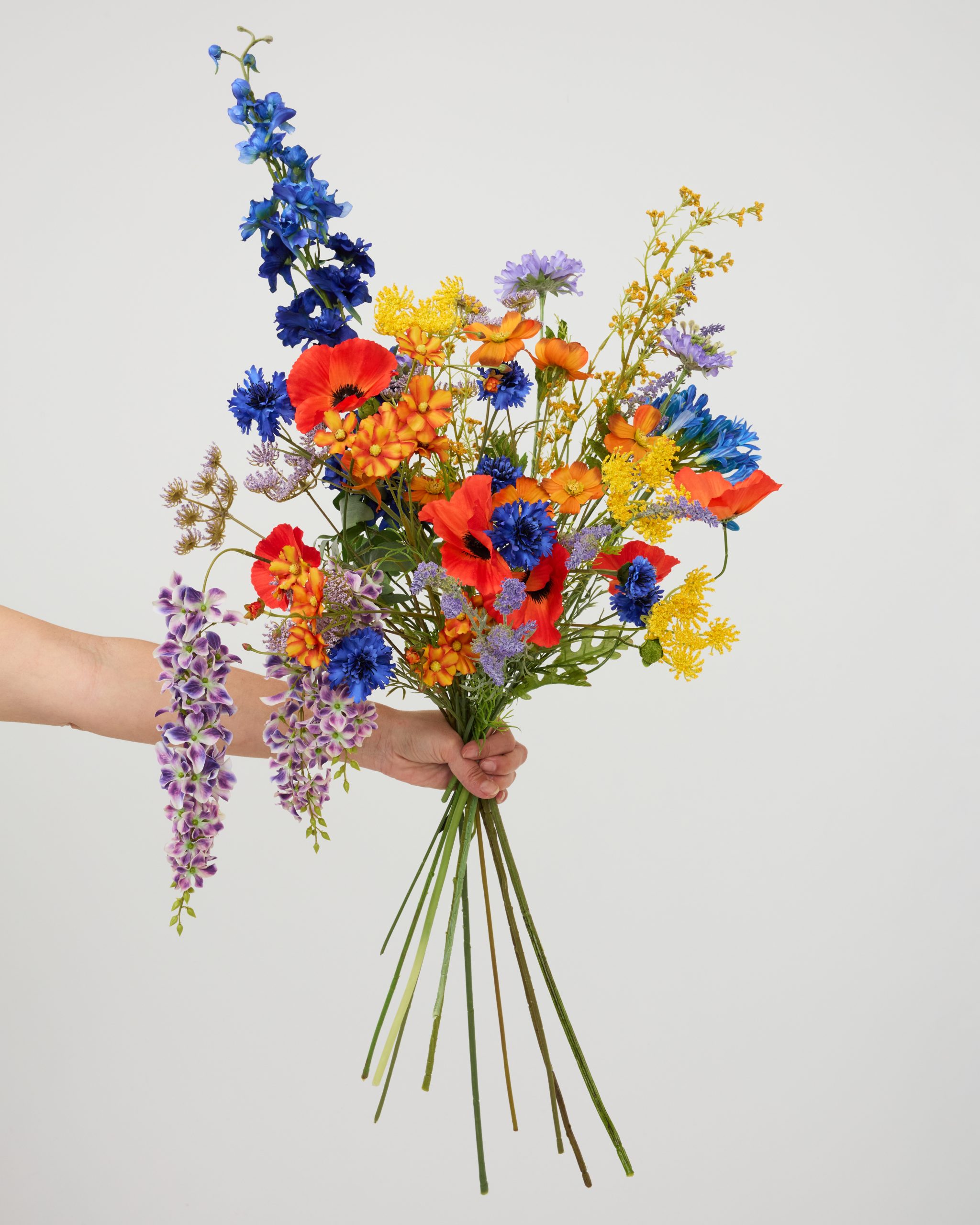 Een hand die een kleurrijk boeket van zijden klaprozen en veldbloemen vasthoudt tegen een lichte achtergrond.