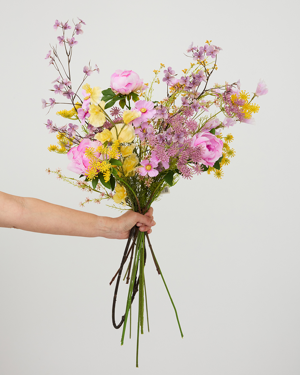 Hand houdt een kleurrijk zijden boeket vast met roze pioenrozen, gele bloemen en fijne paarse accenten tegen een witte achtergrond.