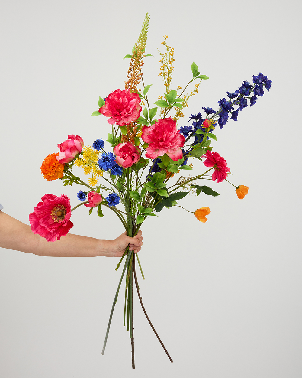 Een hand houdt een kleurrijk boeket van zijden bloemen vast met roze pioenrozen, blauwe riddersporen en oranje accenten.