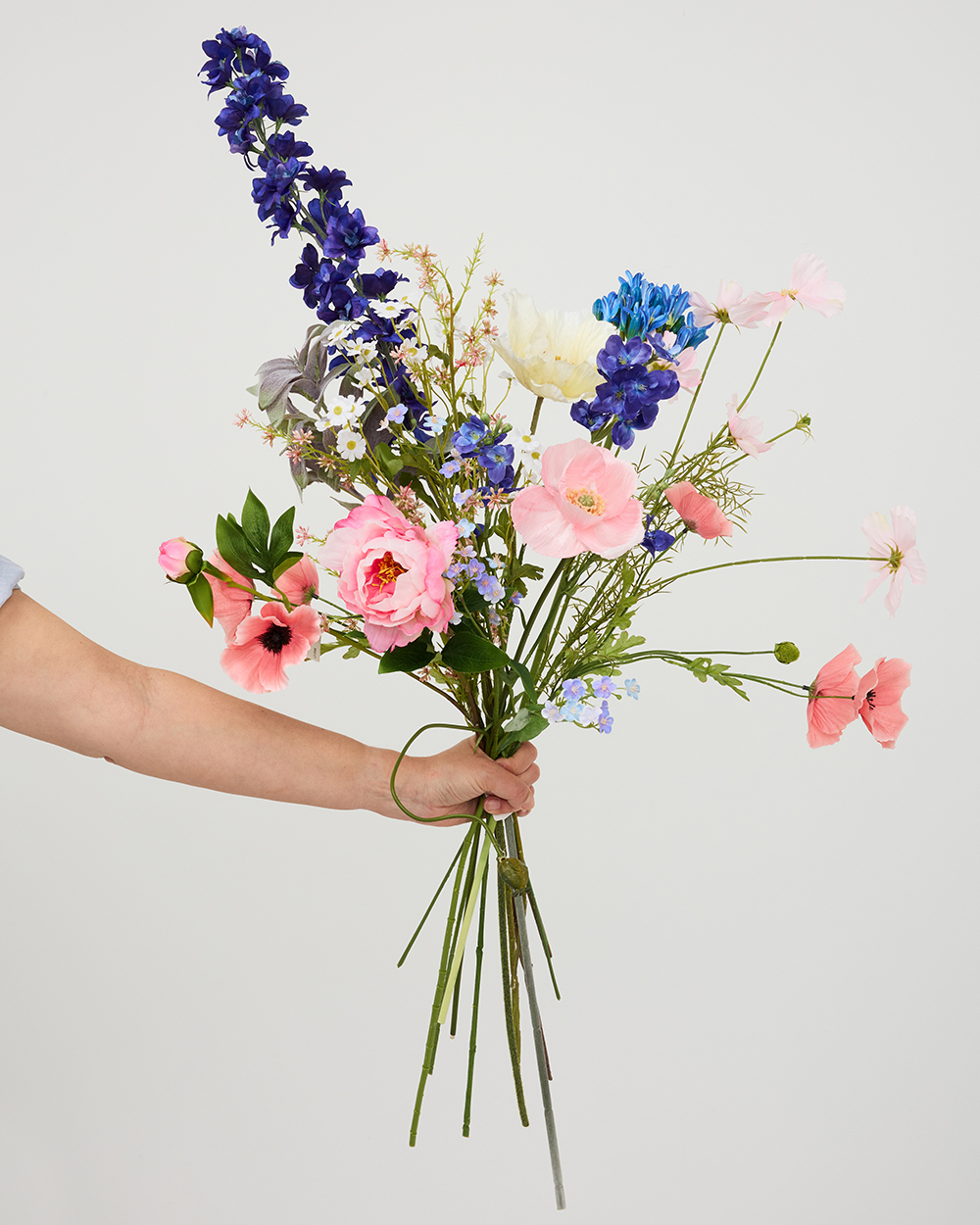 Hand houdt een kleurrijk boeket vast met blauwe ridderspoor, roze pioenen en delicate witte bloemen tegen witte achtergrond.