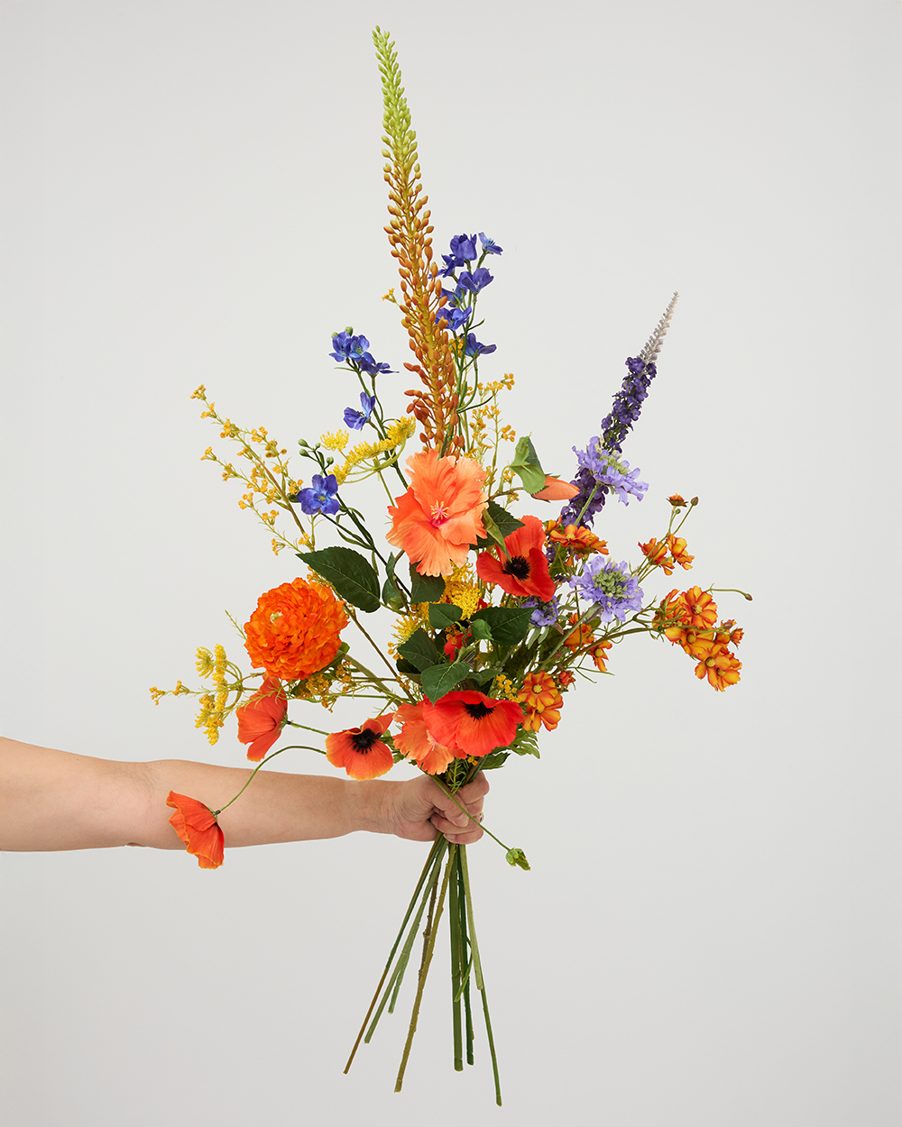 Hand houdt een kleurrijk boeket zijden bloemen vast met oranje, gele en paarse accenten tegen een lichte achtergrond.