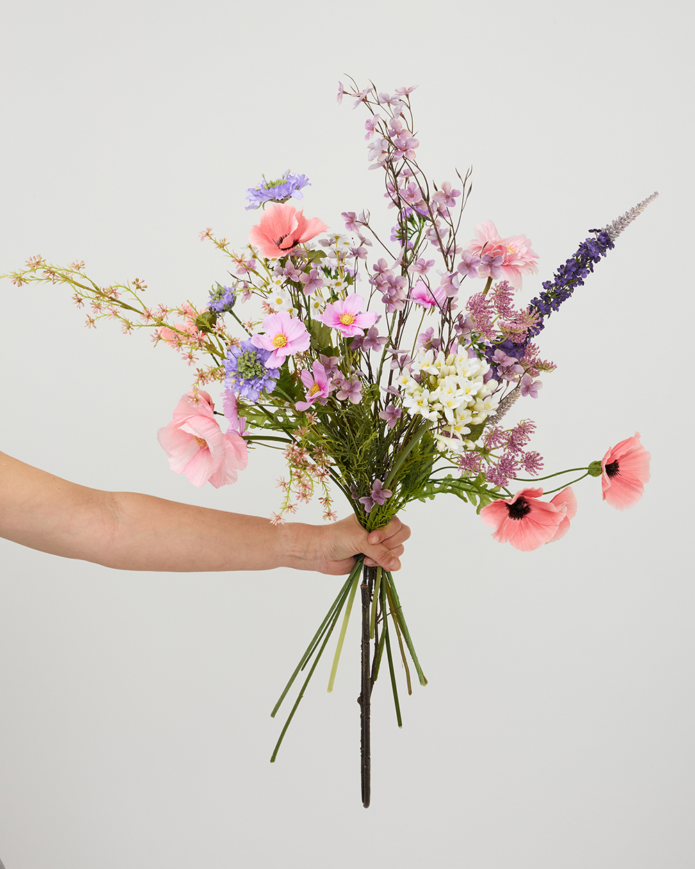 Een hand houdt een elegant veldboeket vast met kunstbloemen in zachte roze, paarse en witte tinten.