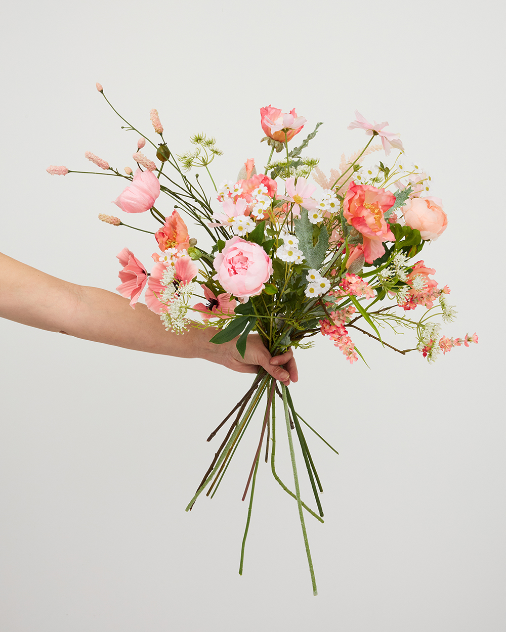 Iemand houdt een handgebonden boeket van zijden bloemen vast in diverse tinten roze, wit en groen tegen een lichte achtergrond.