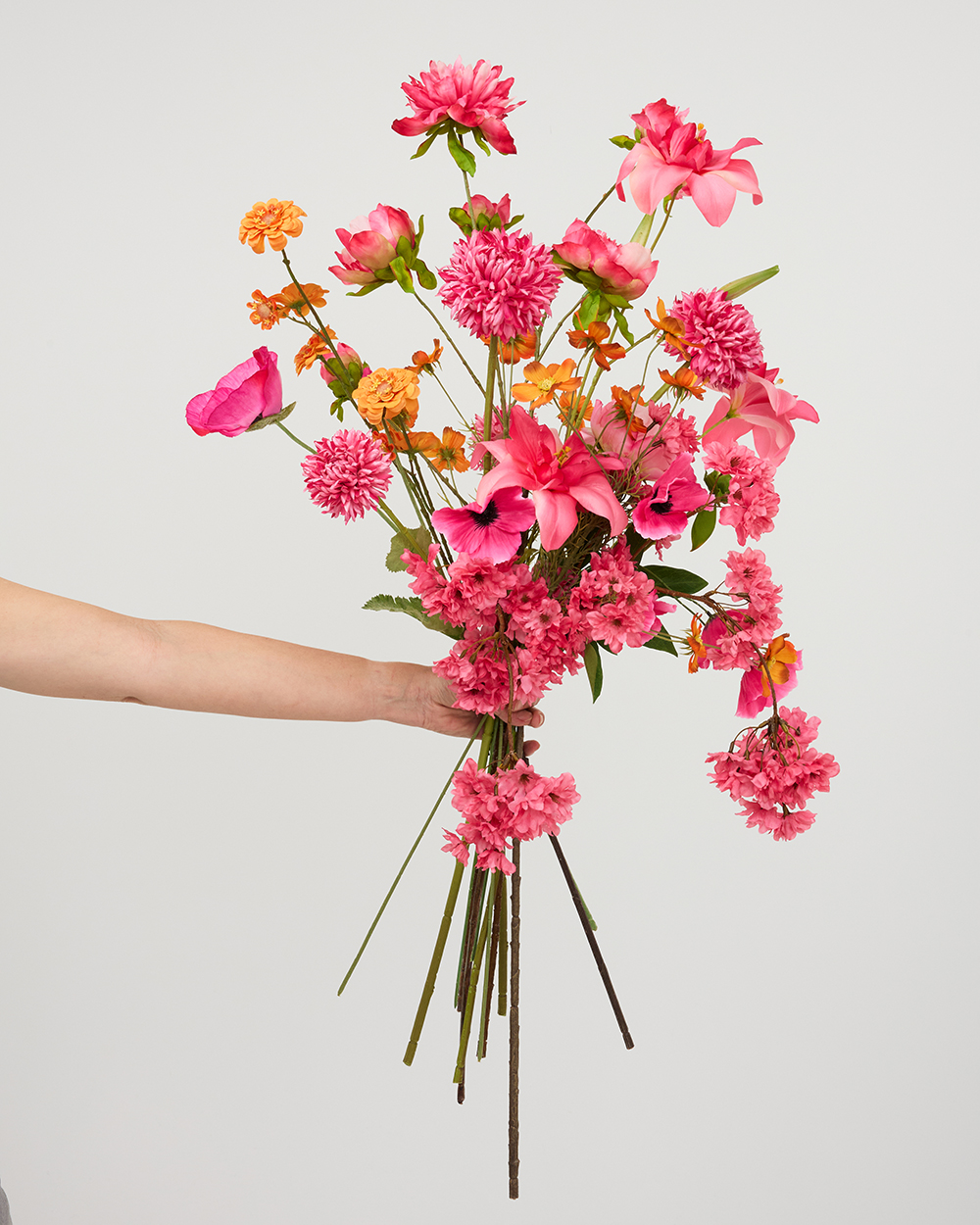 Een hand die een luxueus zijden boeket vasthoudt met roze lelies, dahlia's en oranje accenten tegen een grijze achtergrond.
