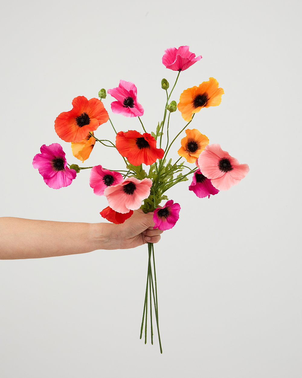 Een hand die een kleurrijk boeket van kunstmatige zijden klaprozen vasthoudt tegen een lichte achtergrond.