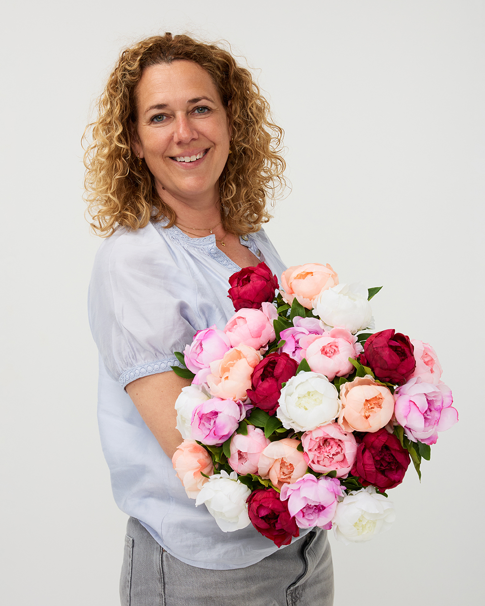 Vrouw met krullend blond haar houdt een weelderig boeket van roze, witte en donkerrode zijden pioenrozen vast.