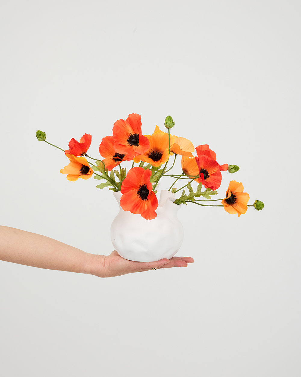 Iemand houdt een witte organische vaas met een boeket zijden oranje en rode klaprozen vast tegen een lichte achtergrond.