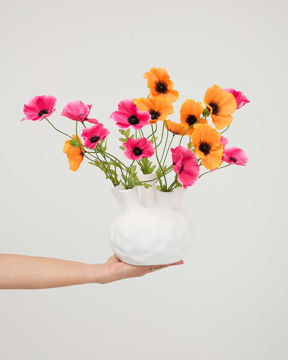 Hand houdt een witte sculpturale vaas vast gevuld met felgekleurde oranje en roze zijden papavers.