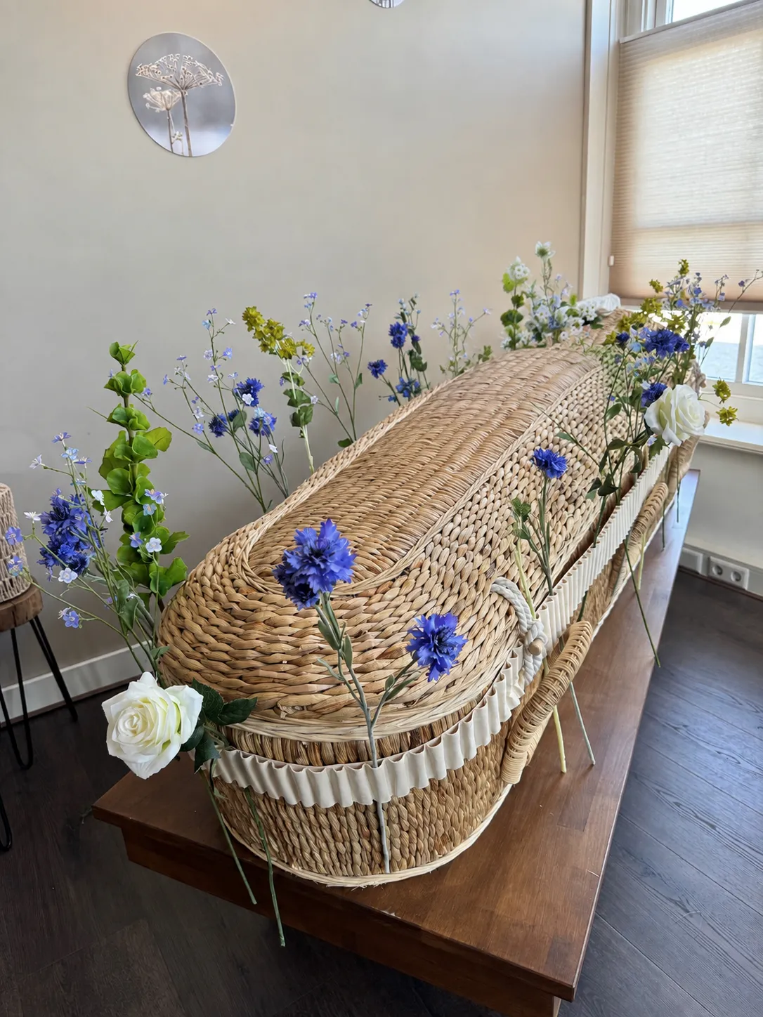 Rieten mand met zijden bloemen in blauw en wit, geplaatst op een houten tafel in een lichte ruimte.