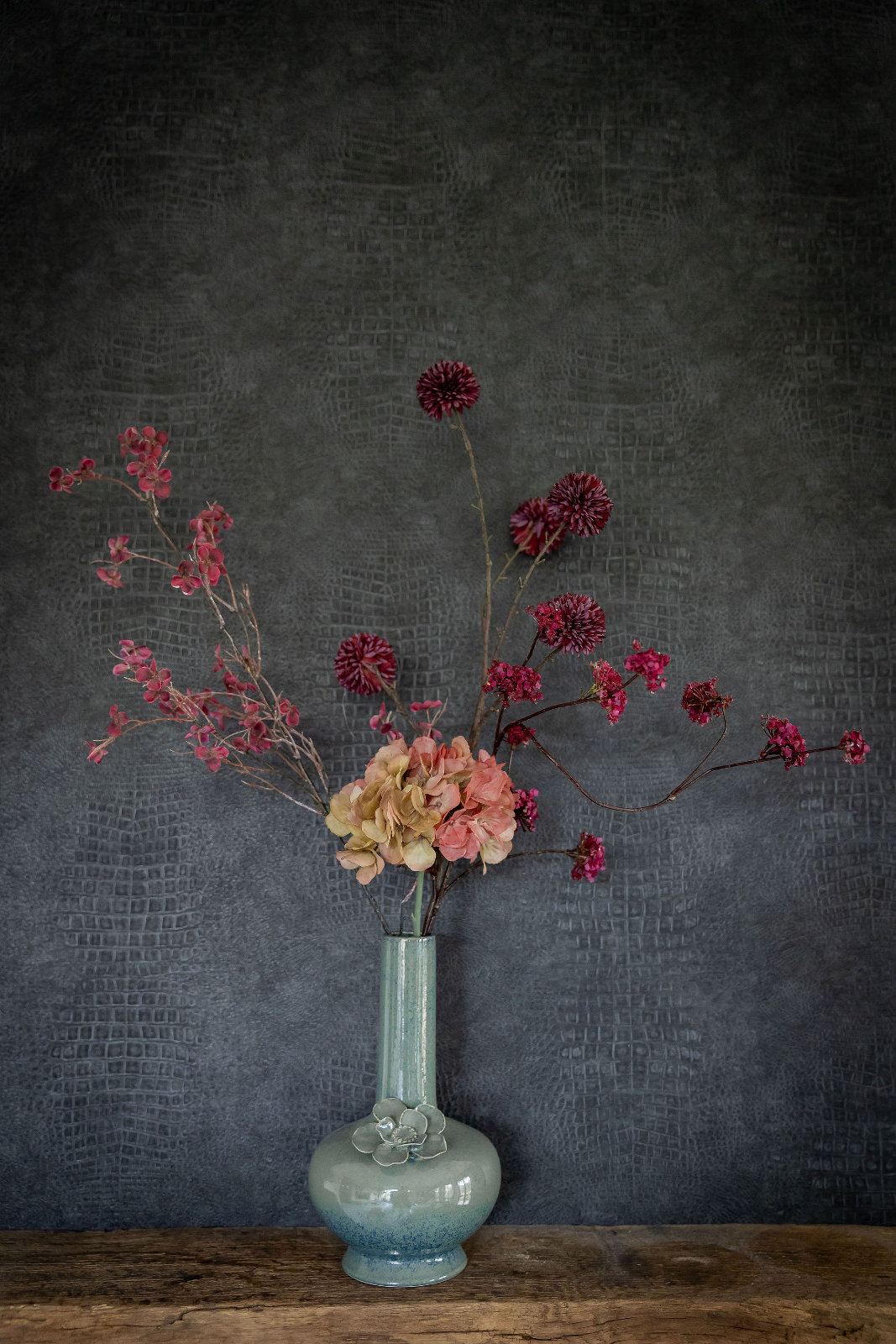 Zachte oranje hortensia, roze bloesemtakken en donkerrode pomponbloemen in een groenblauwe keramische vaas met bloemreliëf op de hals. De vaas staat o