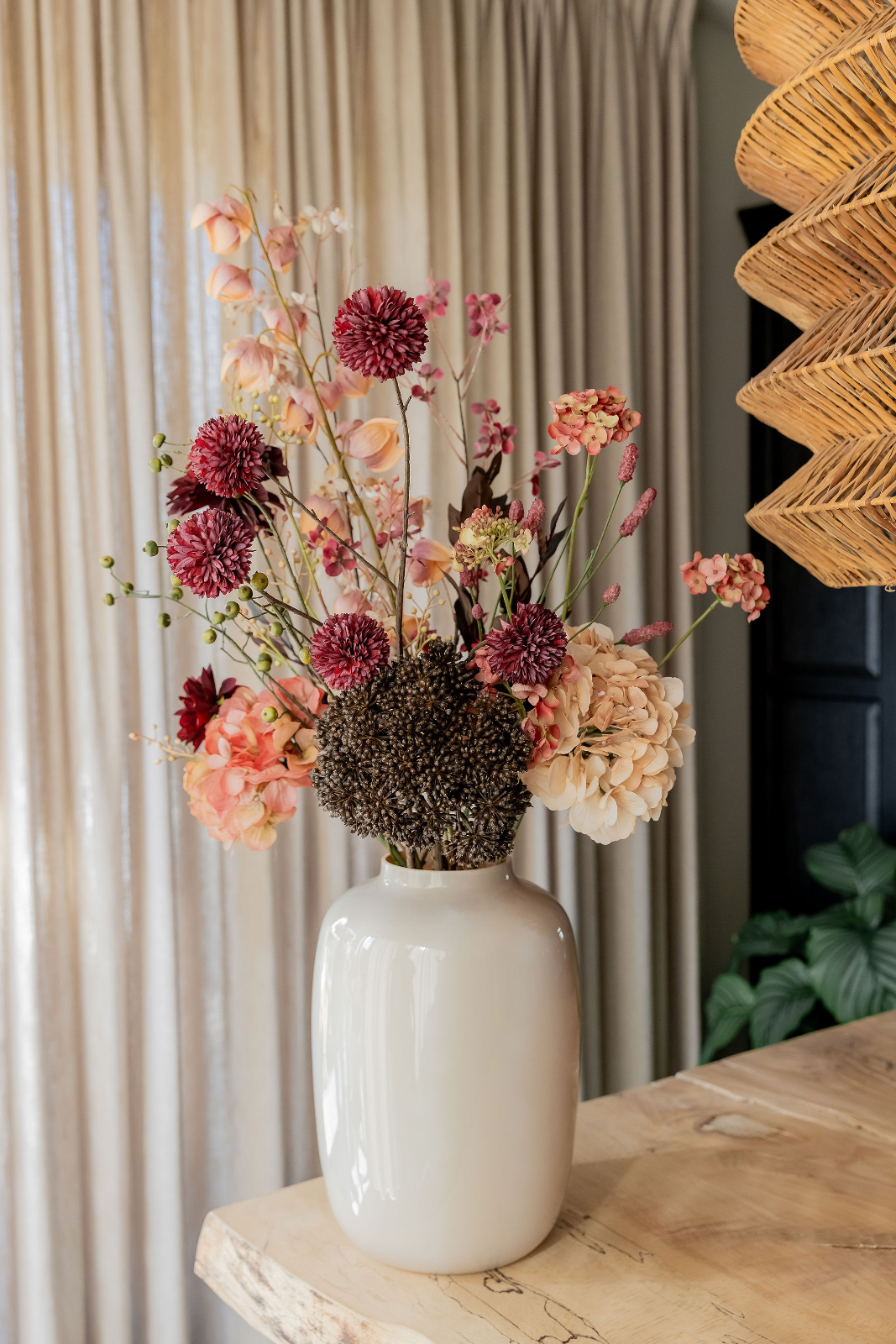 Zijden boeket met donkerrode alliums, perzik- en roze hortensia's, groene besjes en een donkerbruine bolbloem in een witte vaas op een houten tafel, m