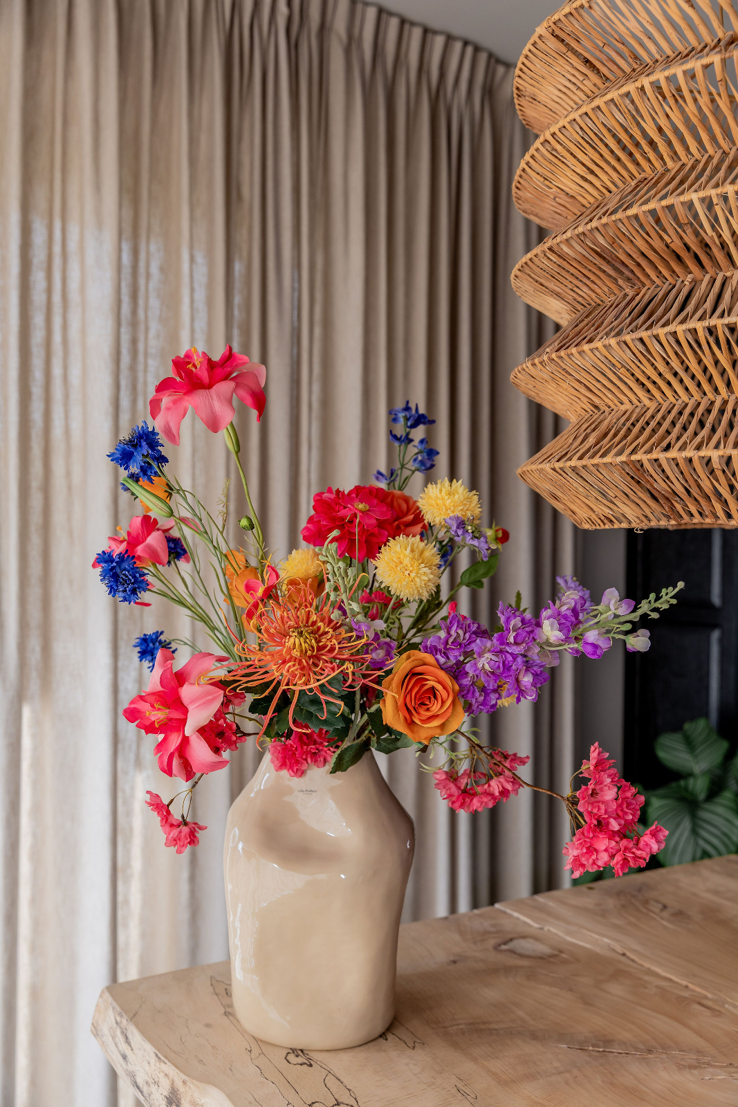 Kleurrijk boeket zijden bloemen in een glanzende beige vaas op een houten tafel, met roze lelies, blauwe korenbloemen, een oranje spinnenchrysant, een