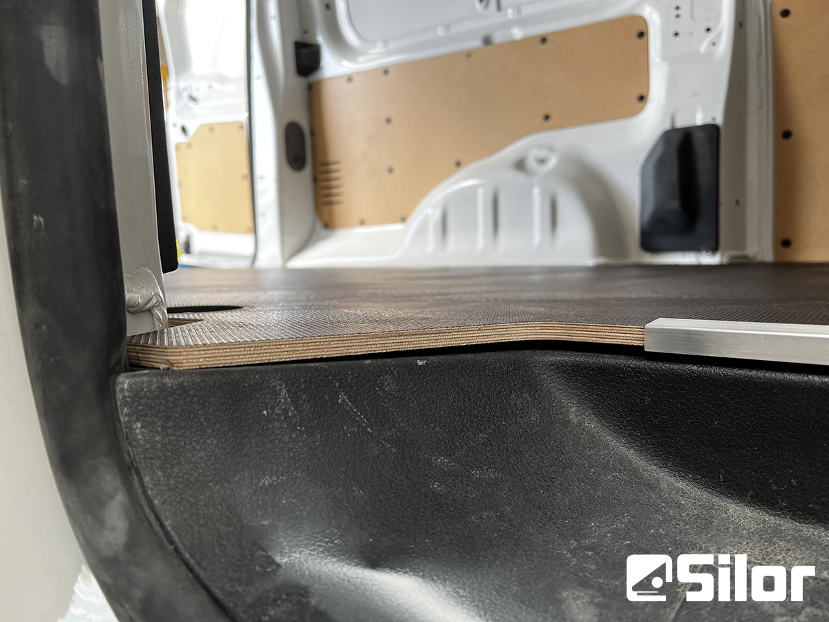 Interno di un furgone con pavimento in legno e pannelli marroni.