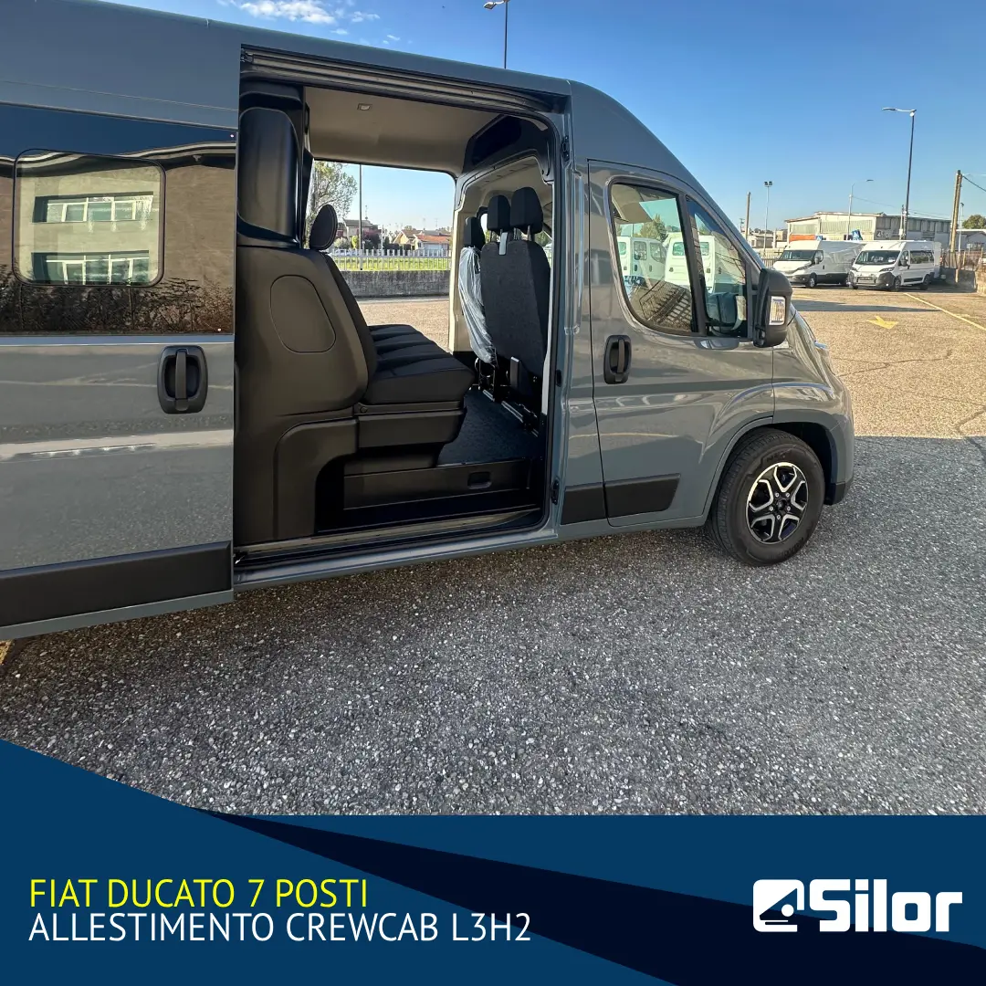 Un furgone grigio chiaro con la portiera scorrevole aperta parcheggiato in un parcheggio con un cielo blu e nuvole sullo sfondo.