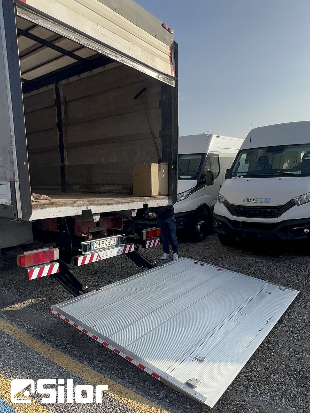 Un camion di consegna bianco con una pedana di sollevamento abbassata è parcheggiato in un parcheggio.