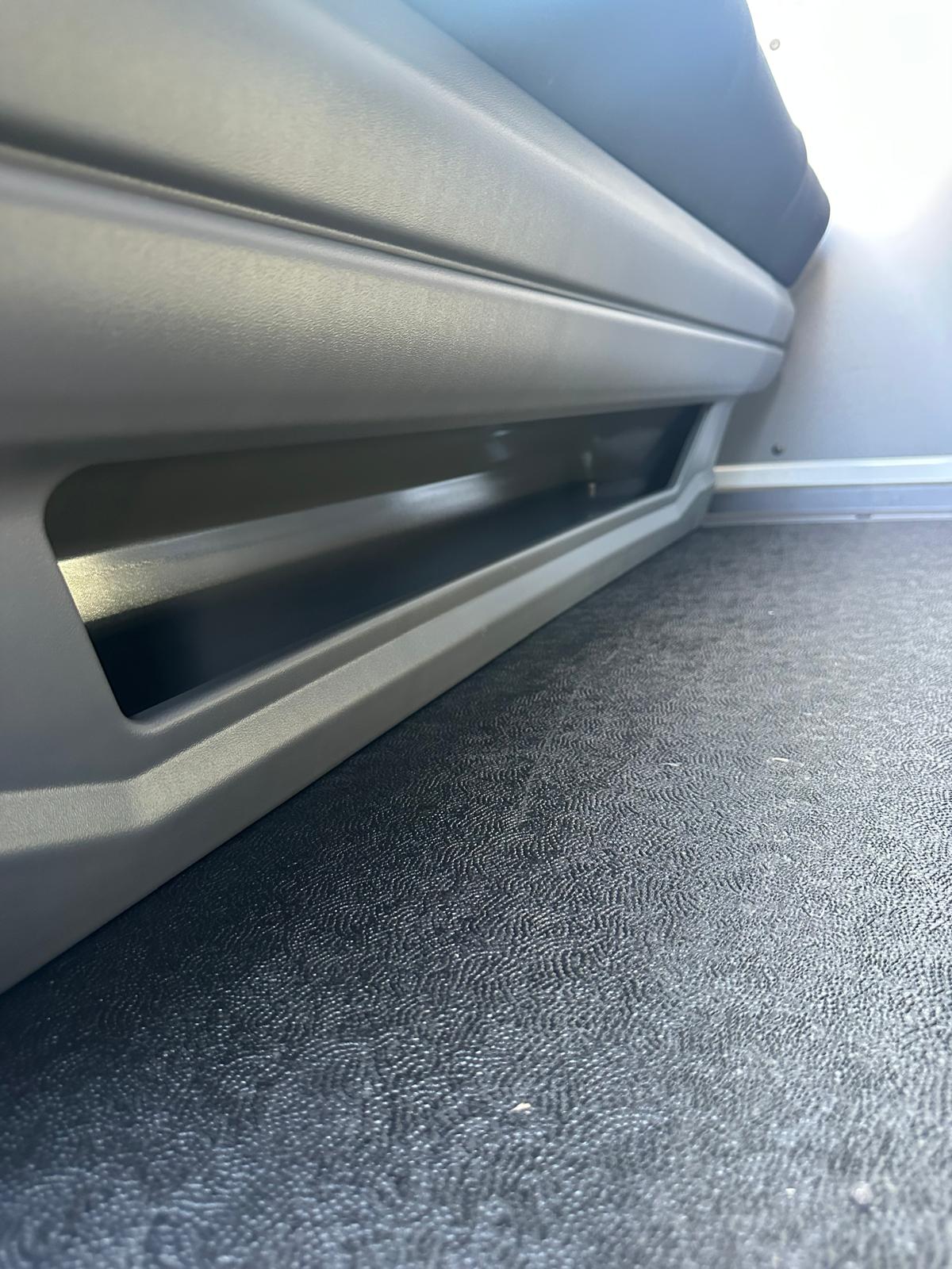 Primo piano di un sedile grigio con un vano portaoggetti sopra un pavimento strutturato nero.