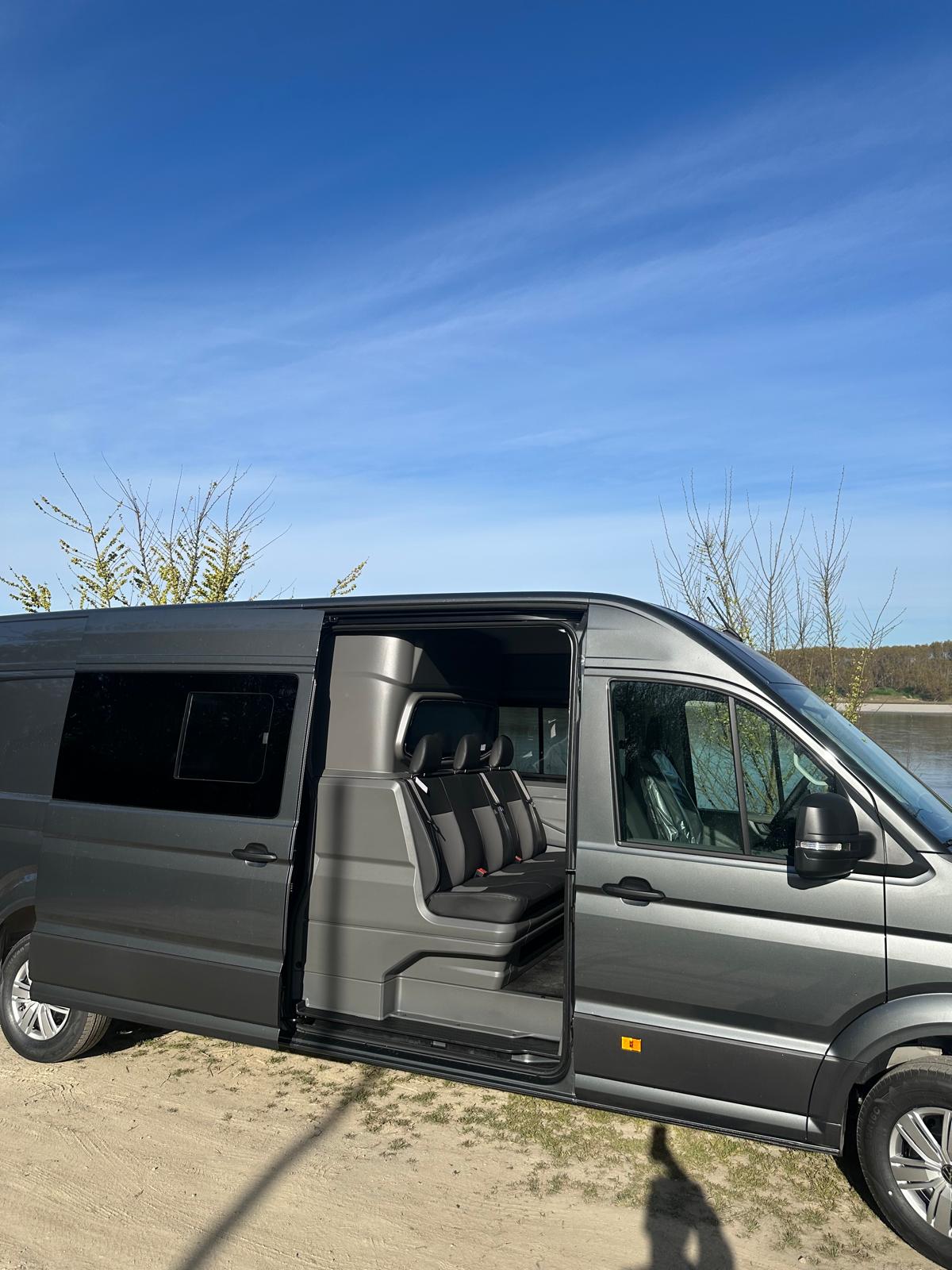 Furgone grigio parcheggiato su una superficie sabbiosa vicino a un lago sotto un cielo azzurro.