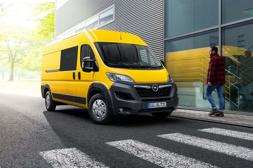Furgone Opel giallo brillante in città: un uomo cammina verso l'edificio moderno.