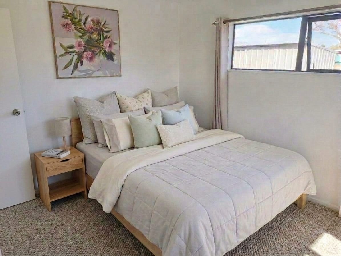 Staged bedroom with layered neutral and pastel cushions, a quilted cover, floral artwork above the bed and a timber bedside table with a lamp