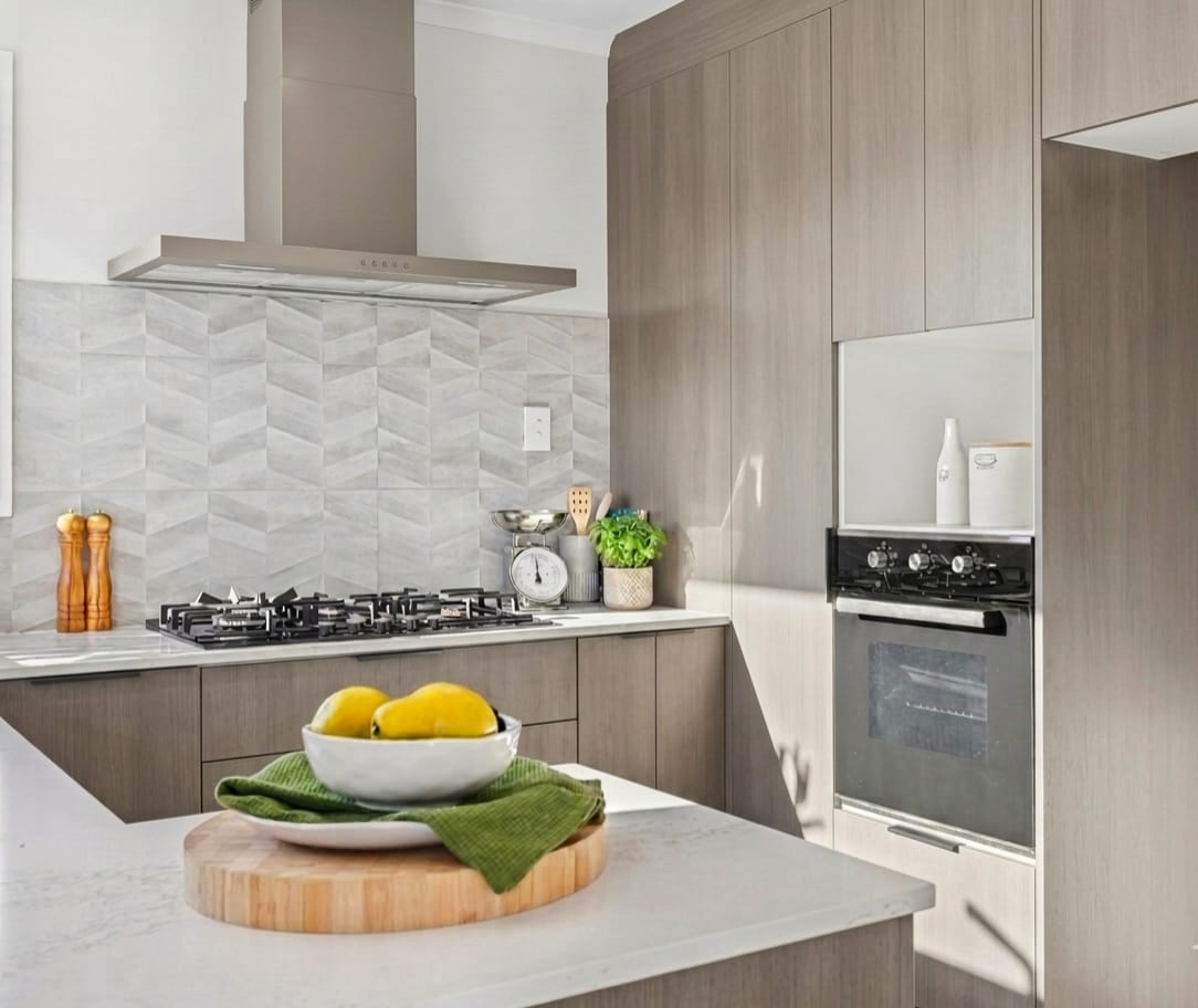 Kitchen with timber-look cabinetry, patterned tile splashback, stone benchtops and a bowl of lemons on a round timber board