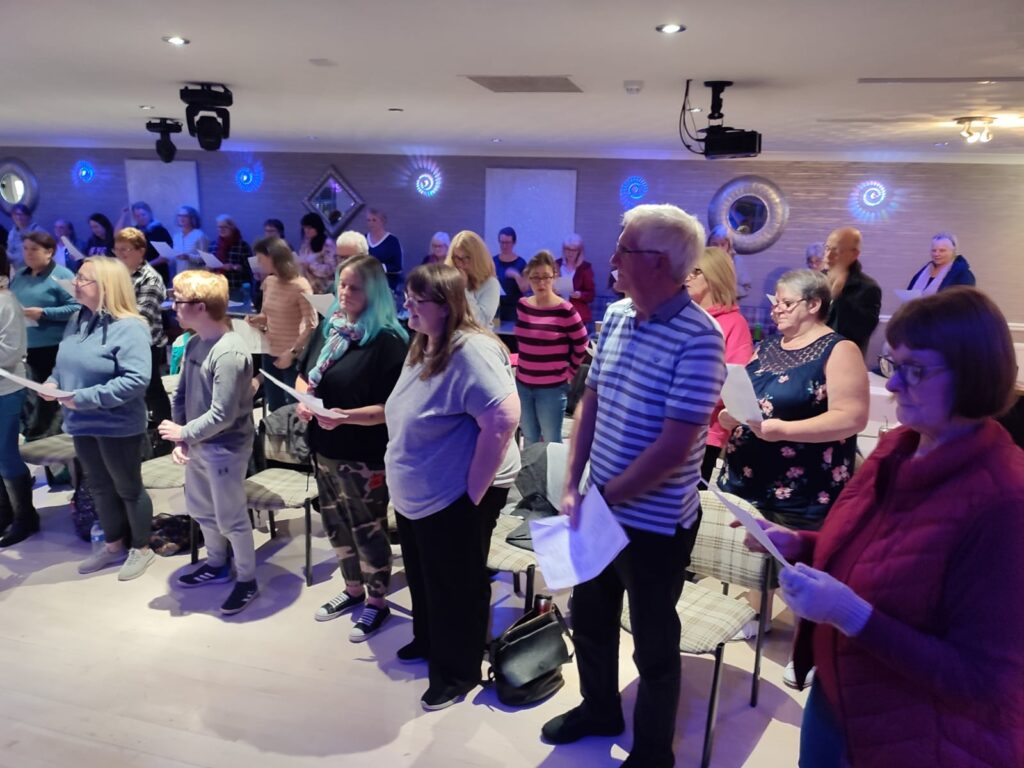 The choir standing on stage mid-performance, warmly lit, smiling as they sing