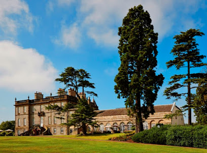 Historic stone country house on landscaped lawns, framed by tall trees under a blue sky.