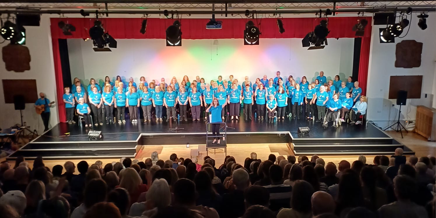 Full Singergie Dunfermline choir, on stage, in front of audience, with conductor facing audience, and guitarist to the side.