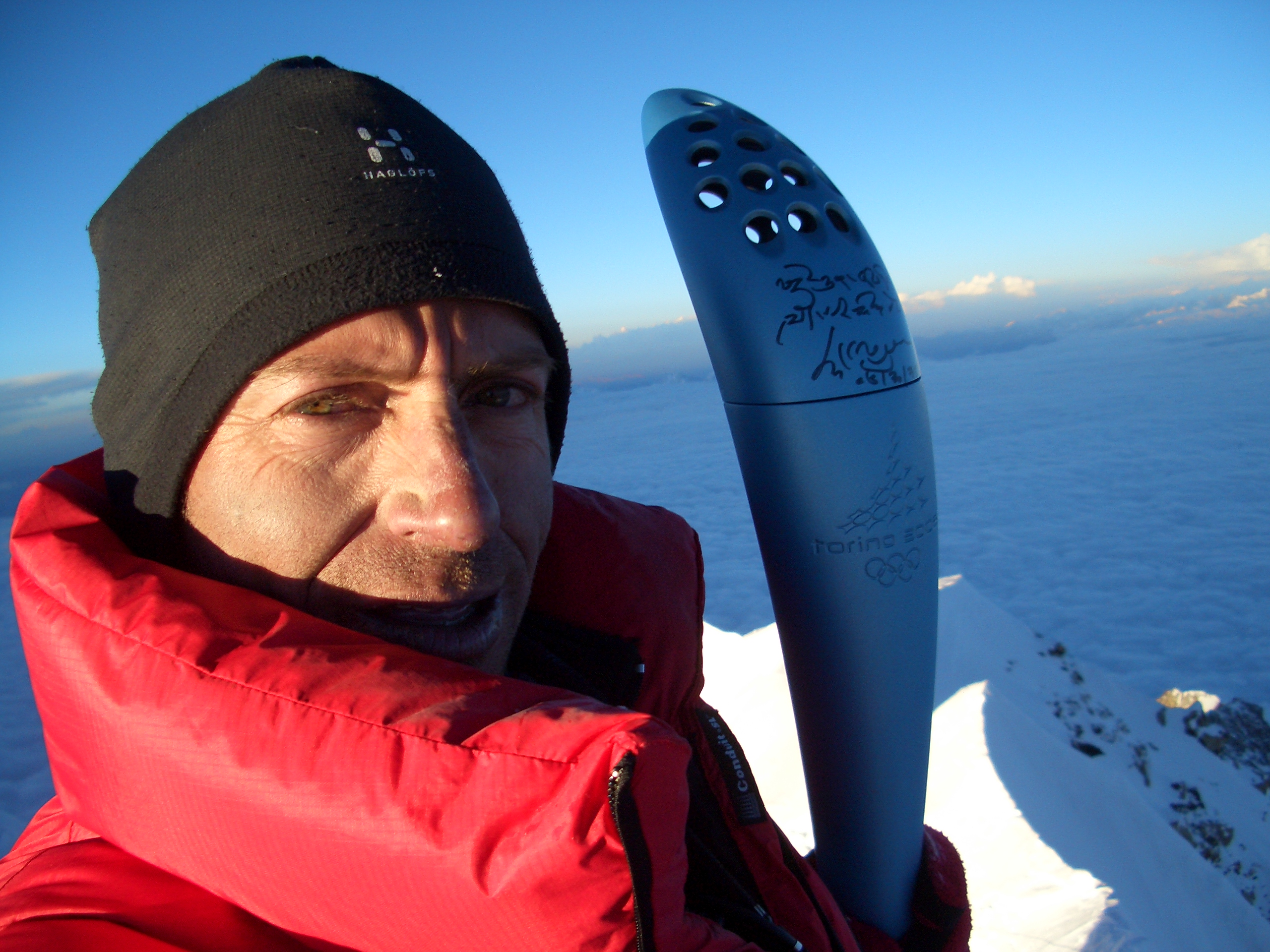 Uomo che detiene la torcia olimpica Torino 2006 in cima a una montagna innevata.